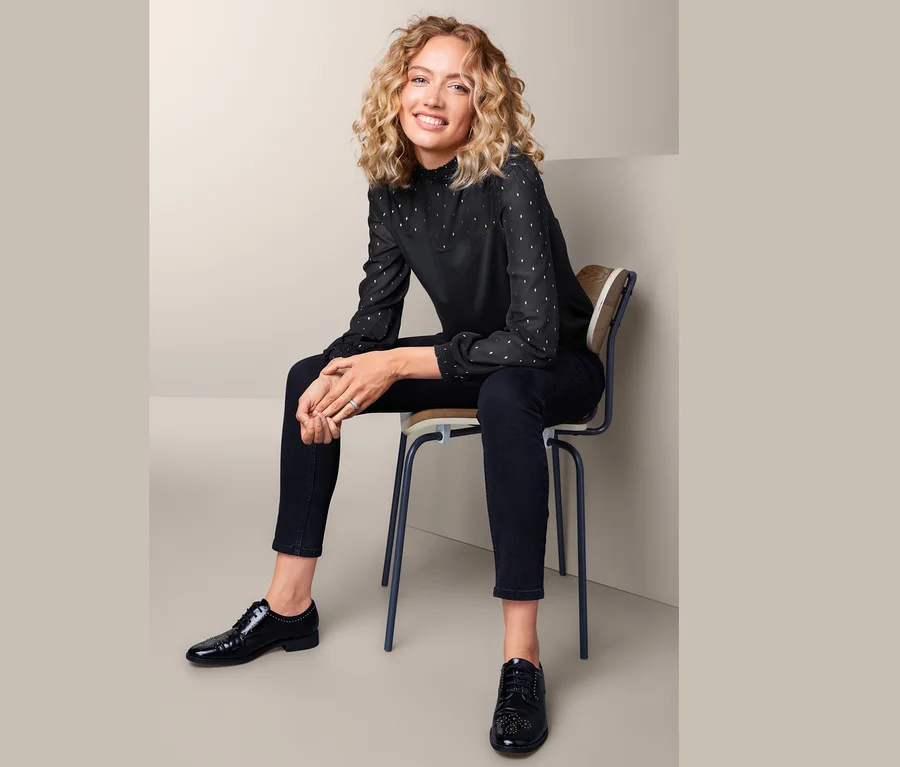 Frau sitzt auf einem Stuhl und trägt ein Blusenshirt im Materialmix, schwarze Hose und schwarze Schuhe.