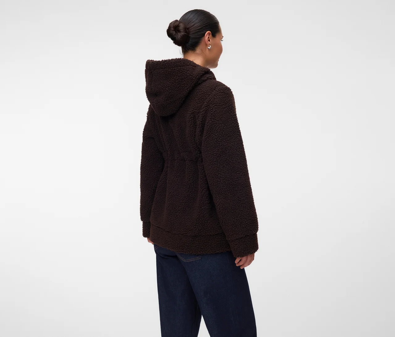 Frau von hinten gesehen, trägt eine dunkelbraune Kapuzenjacke aus Teddyfell mit Kordelzug in der Taille und dunkelblaue Jeans. Ihr Haar ist zu einem Dutt gebunden, und sie trägt kleine Creolen.