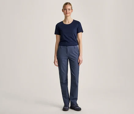 Eine junge Frau steht frontal vor einem hellen Hintergrund, trägt ein dunkelblaues T-Shirt, eine blaue Hose und dunkelblaue Schuhe.