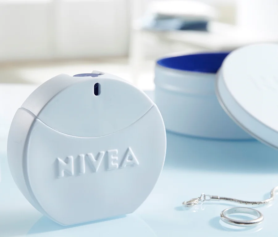 Ein hellblaues, rundes NIVEA Produkt, möglicherweise ein Parfüm oder Deodorant, steht auf einer hellblauen Oberfläche neben einem silbernen Ring und einer Kette. Im Hintergrund ist eine offene NIVEA Cremetiegel mit blauem Inneren zu sehen.