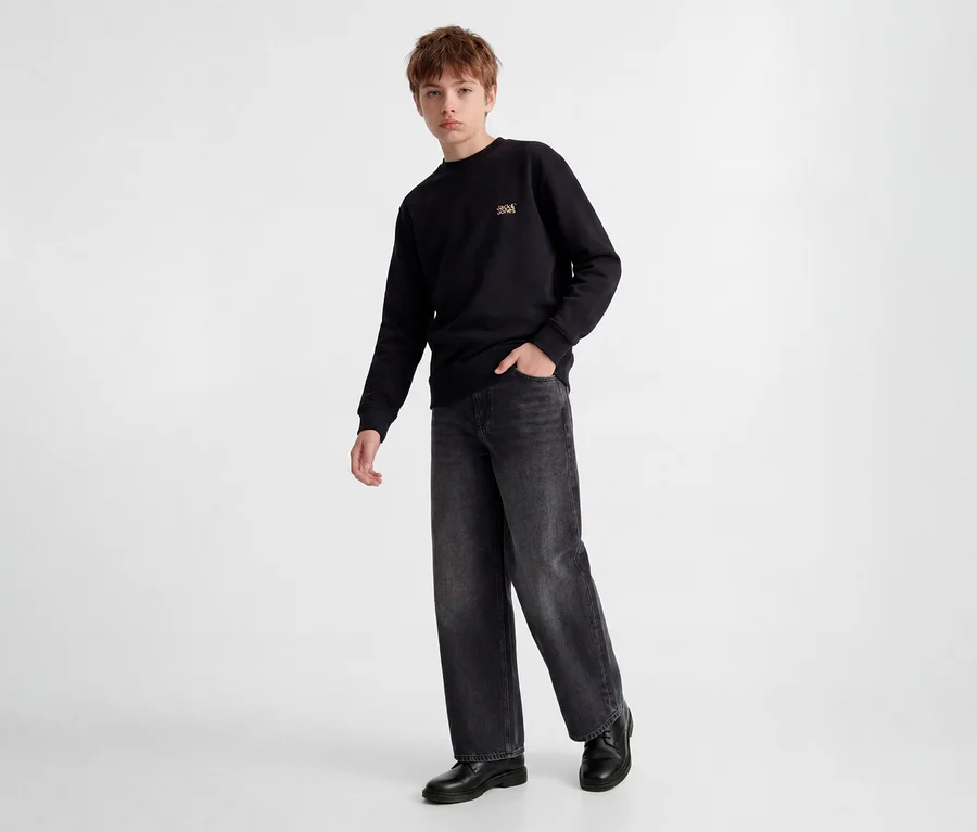 Junger Junge steht in schwarzem Sweatshirt mit kleinem goldenem Logo, dunkelgrauen Wide-Leg Jeans und schwarzen Schuhen. Eine Hand steckt in der Hosentasche.