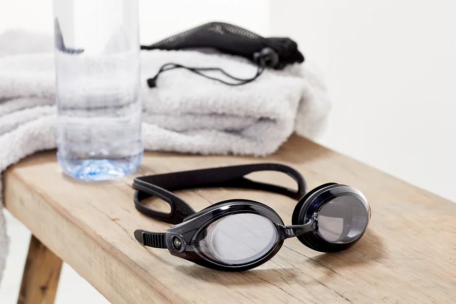 Schwarze Schwimmbrille liegt auf einer Holzbank neben einer Wasserflasche und einem gefalteten weißen Handtuch.