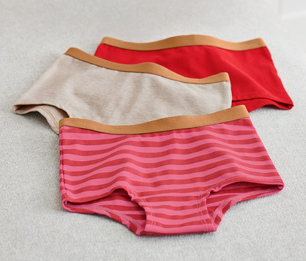 Auf einer hellgrauen Oberfläche liegen drei Kinder-Pantys, eine beige, eine rote und eine rosa gestreifte. Links davon liegt eine rosa Decke.