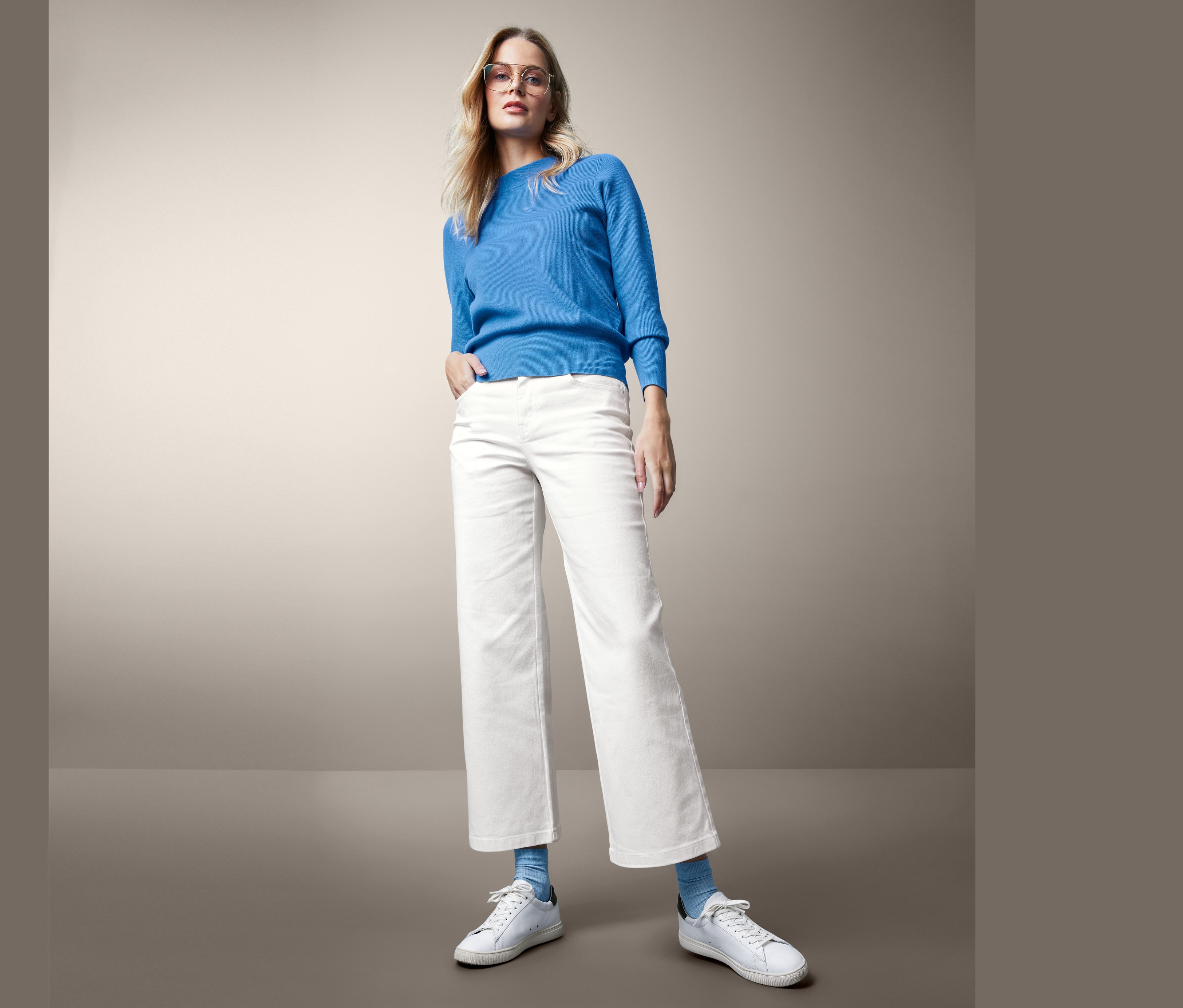 Blonde Frau posiert in weissen Culotte-Jeans und blauem Pullover.