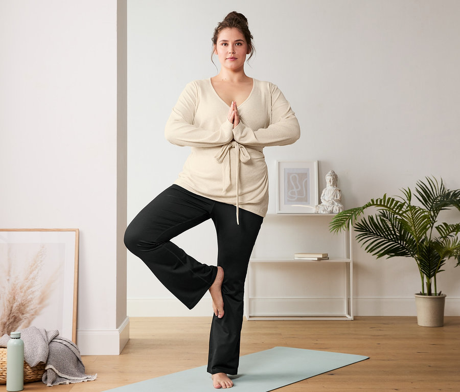 Eine Frau in Yoga-Wickel-Cardigan steht in der Baum-Pose auf einer Yogamatte und trägt eine schwarze Bodyforming-Sporthose.