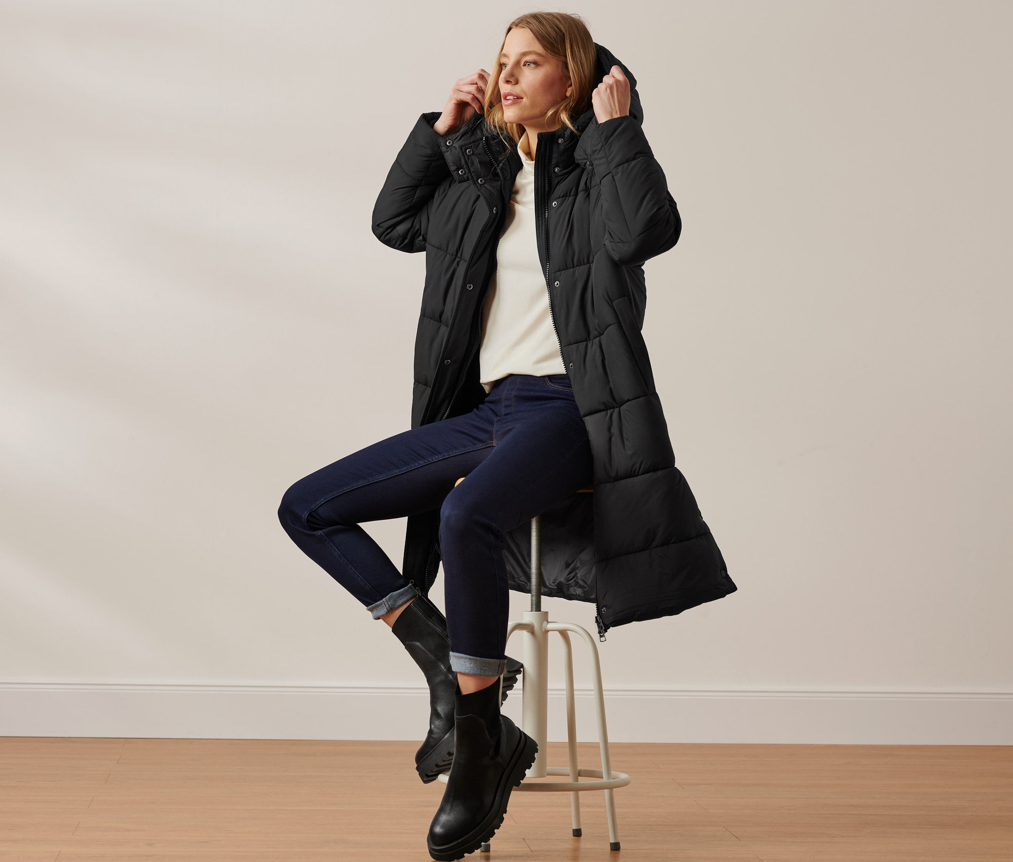 Frau sitzt auf einem Hocker und trägt einen schwarzen Steppmantel mit Kapuze.