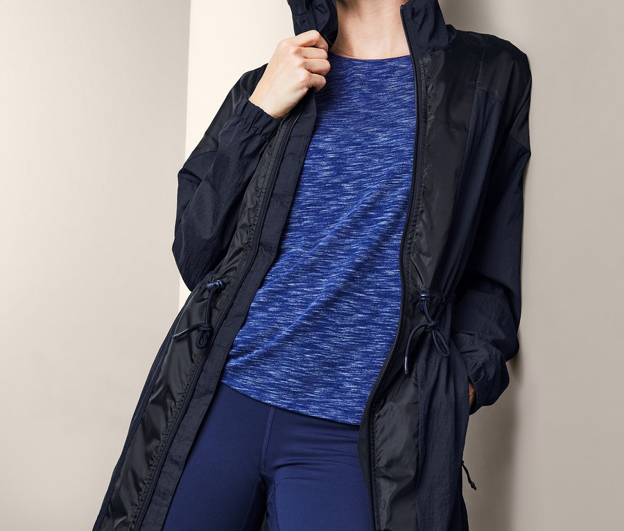 Blonde Frau lehnt an einer Wand und trägt eine Funktionsjacke, ein blaues T-Shirt und Leggings.