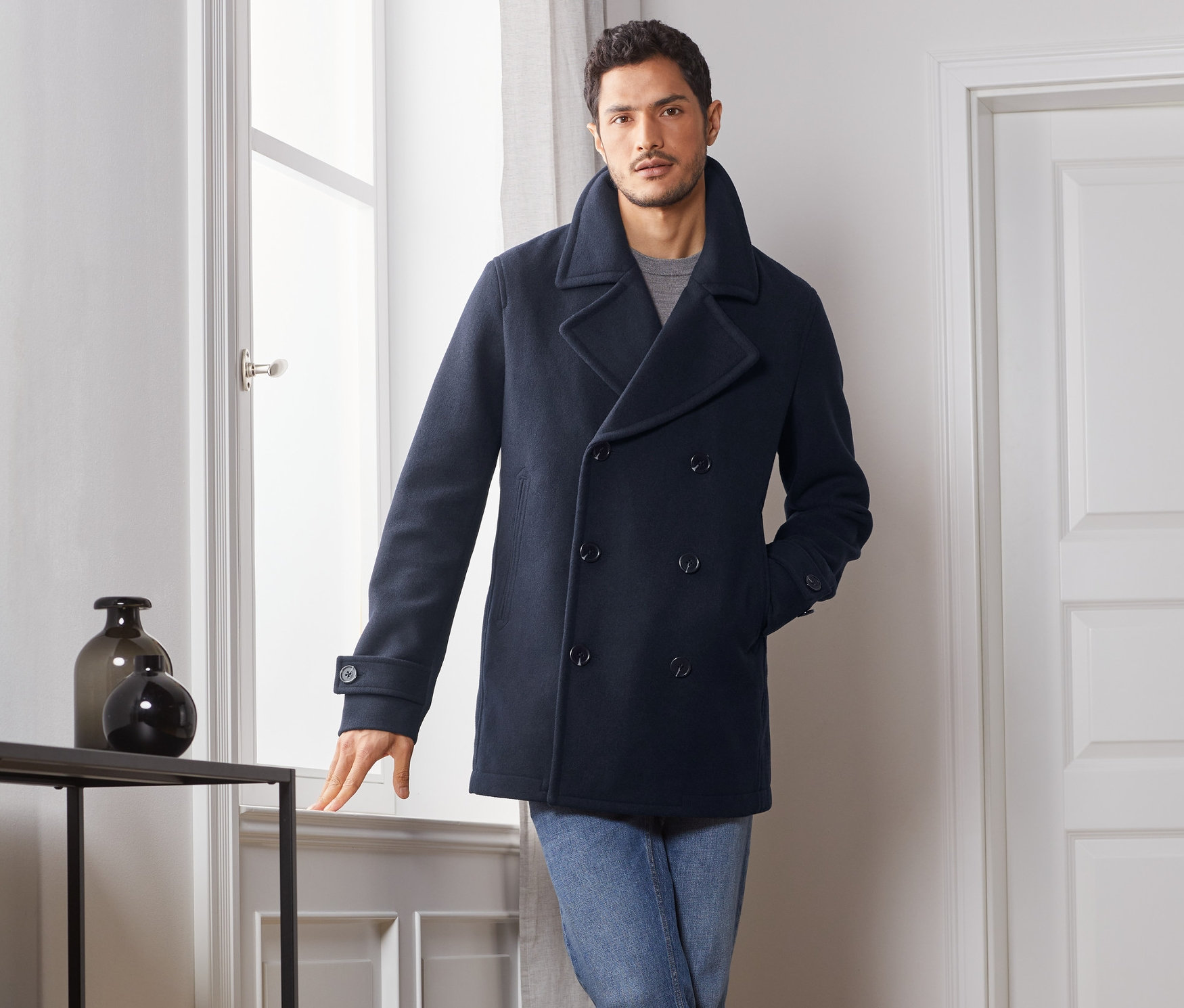 Mann posiert am Fenster in Caban-Jacke, Jeans und Stiefeln.