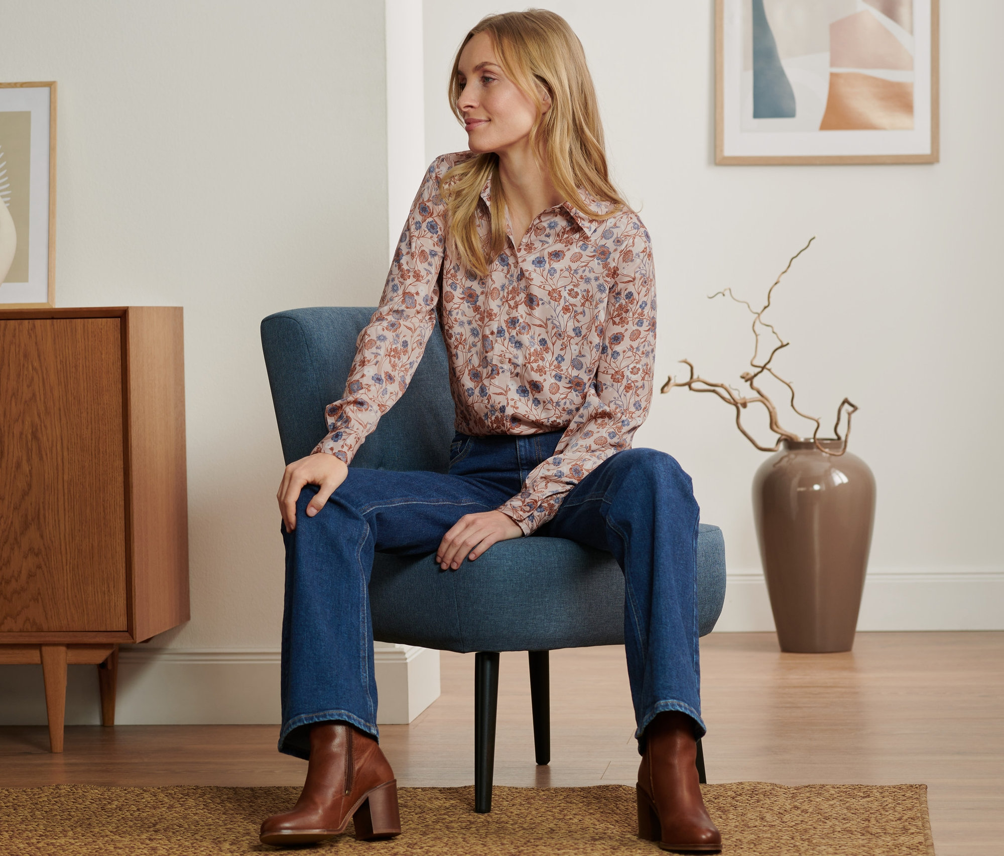 Frau sitzt im Sessel und trägt eine florale Bluse, Jeans und braune Stiefel.