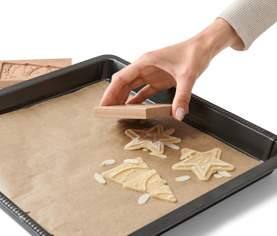 Eine Hand drückt eine hölzerne 2 Spekulatiusformen auf sternförmigen Teig auf einem Backblech mit Backpapier.