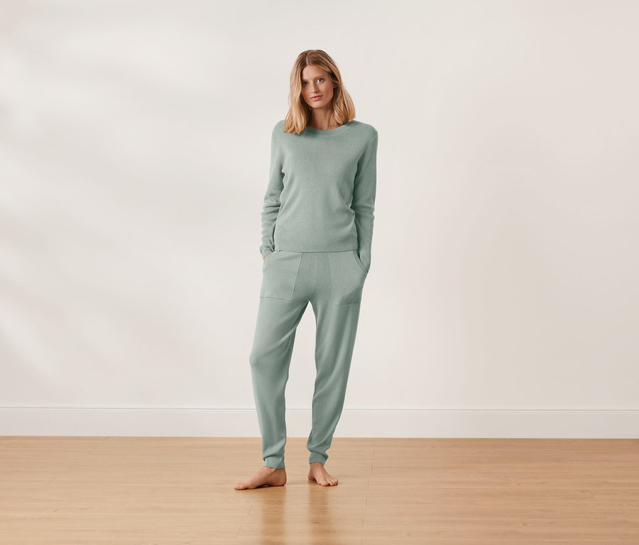 Frau steht barfuß und trägt einen grünen Hausanzug aus Strick.