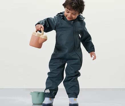 Ein Kind im Kleinkinder-Regeneinteiler hält eine Gießkanne und steht neben einem Eimer.