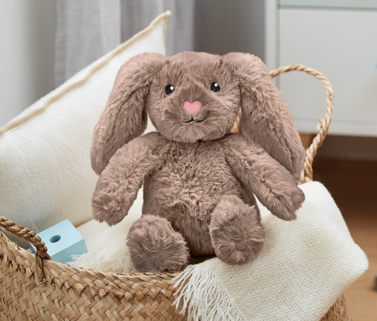 Ein Plüsch-Hase sitzt in einem Weidenkorb auf einer weißen Decke neben Holzbausteinen.