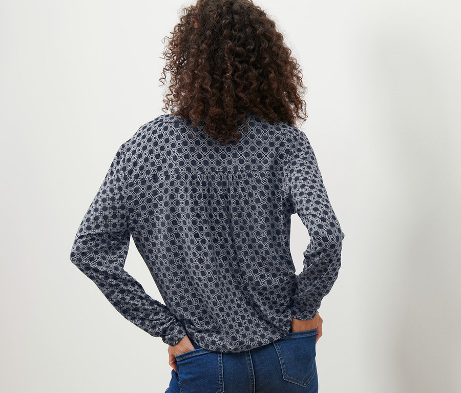 Rückansicht einer Frau mit Händen in den Hosentaschen, die ein bedrucktes Blusenshirt trägt.