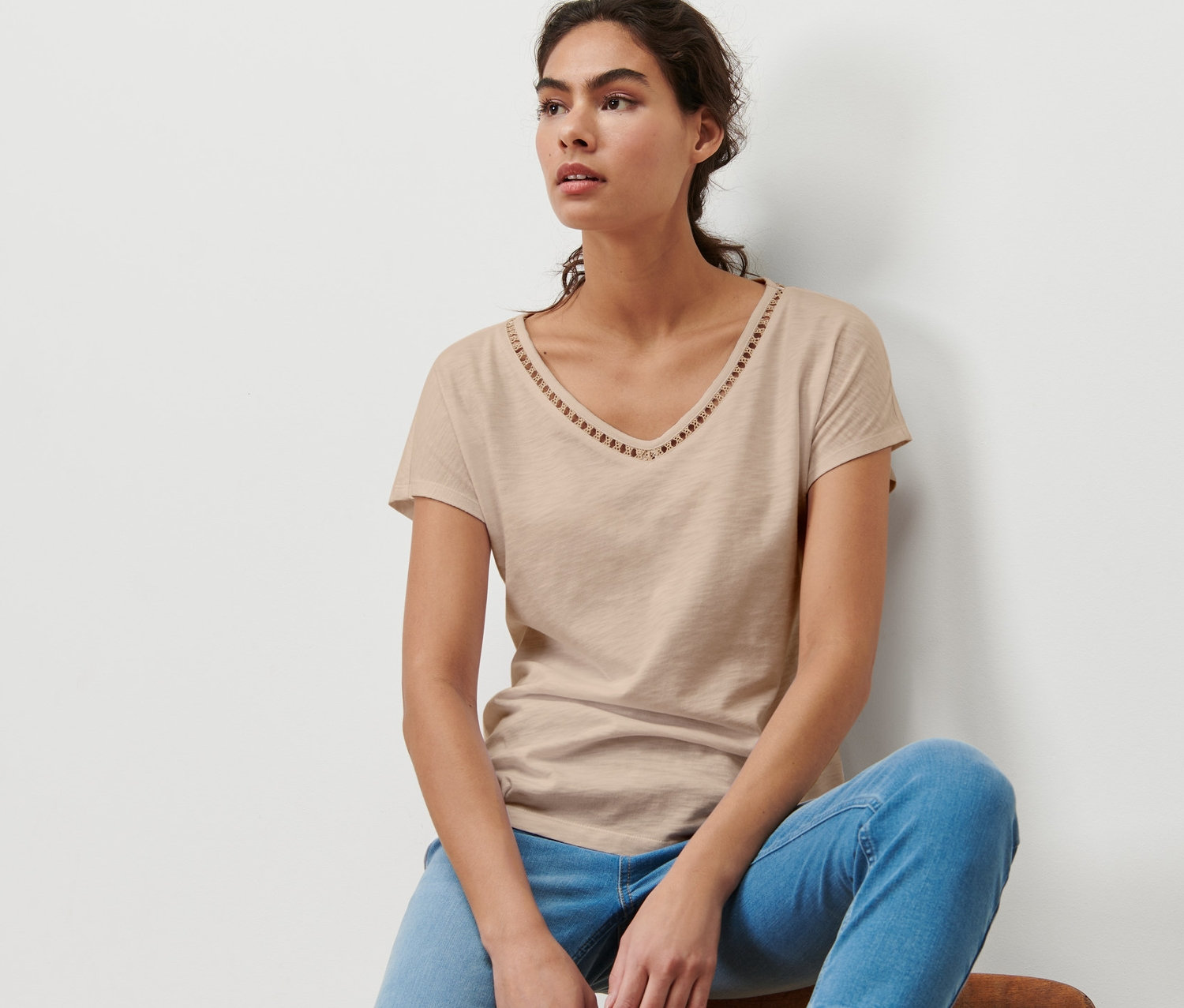 Eine Frau sitzt auf einem Stuhl und trägt ein beiges Shirt mit Crochet-Tape und blaue Jeans.