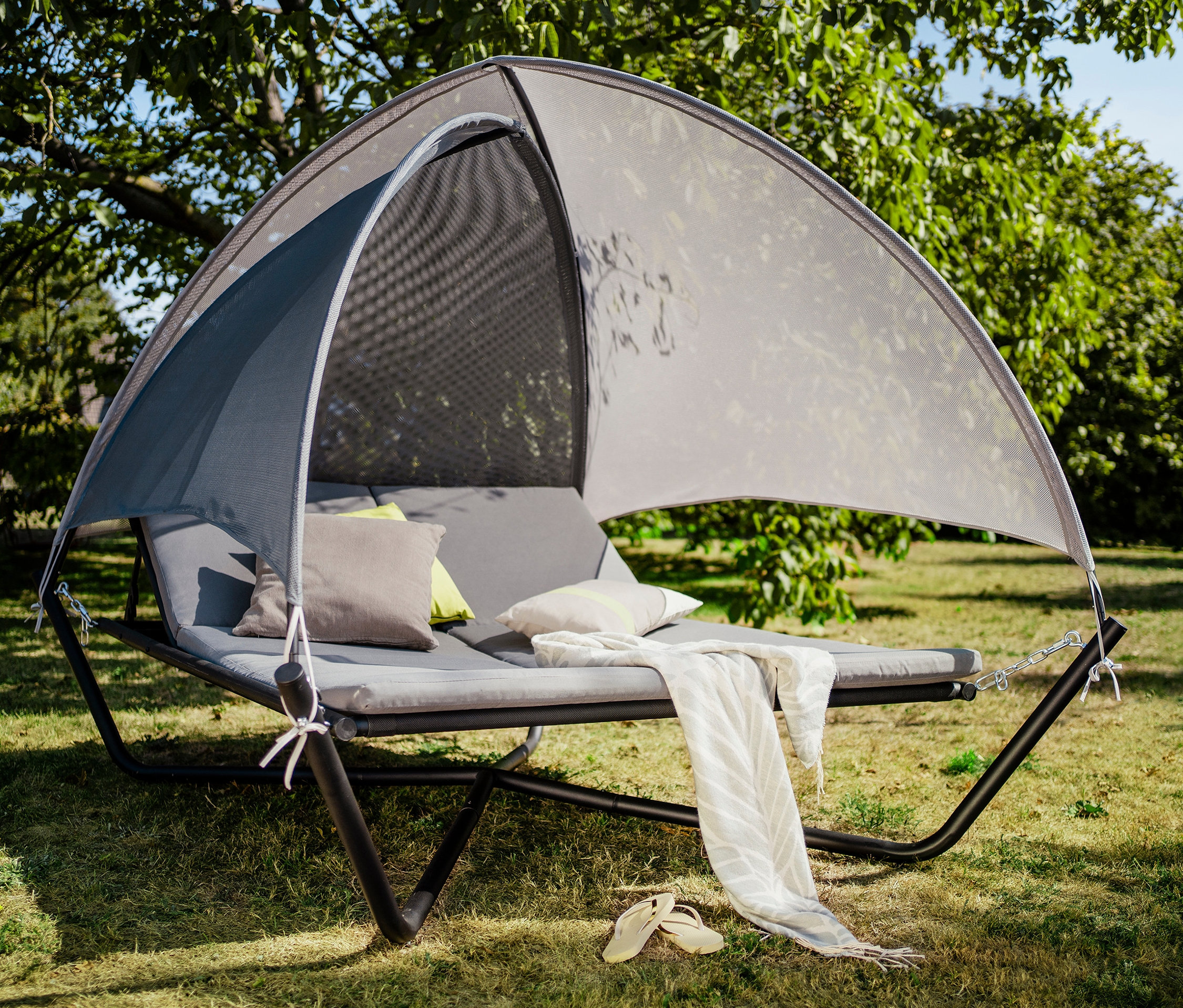 Graue Sonnenliege mit Baldachin, Kissen und Überwurf auf einer Rasenfläche.