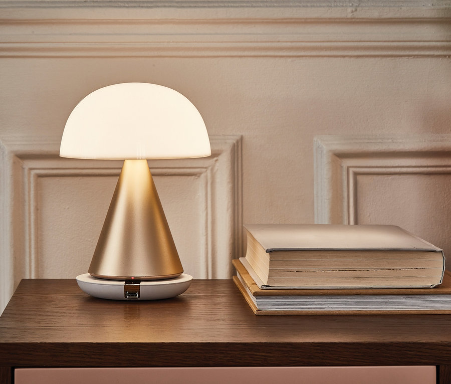 Auf einem braunen Schrank steht eine goldene LED-Lampe Lexon Mina L neben Büchern und einem Blumentopf.