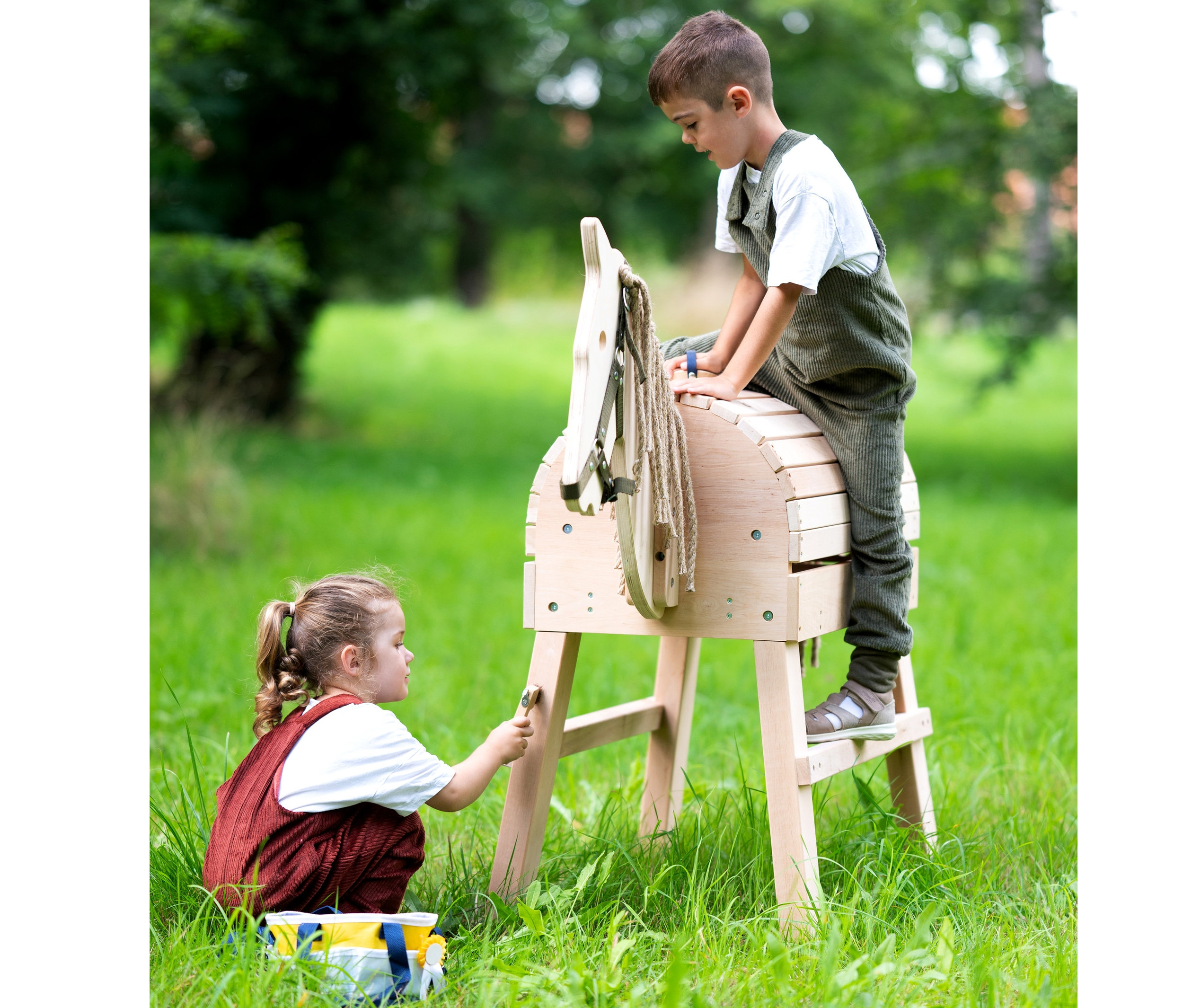 Ein Junge sitzt auf einem Holzpferd, ein Mädchen steht daneben und spielt damit. Am Pferd ist ein Zaumzeug befestigt. Im Gras steht eine Tasche mit einem small foot Spielzeug-Pferdepflege-Set mit Tasche.
