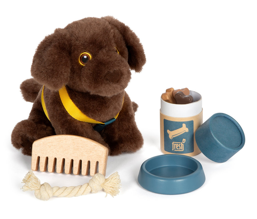 Ein brauner small foot Plüschhund mit gelbem Geschirr sitzt neben einem Kamm, einem Kauspielzeug, einem Napf und einem Behälter mit Leckerlis.