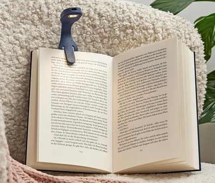 Eine blaue LED-Mini-Leselampe mit Akku ist an einem offenen Buch befestigt, das auf einem Sessel liegt.