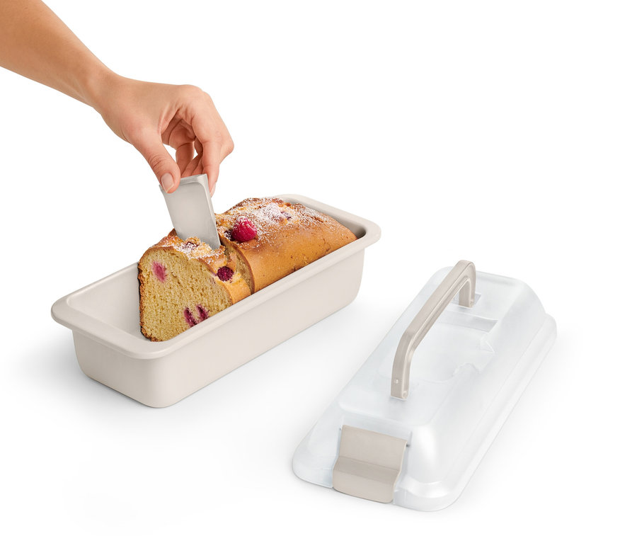 Eine Hand schneidet ein Stück Himbeerkuchen in einer Kastenform mit Transportdeckel.