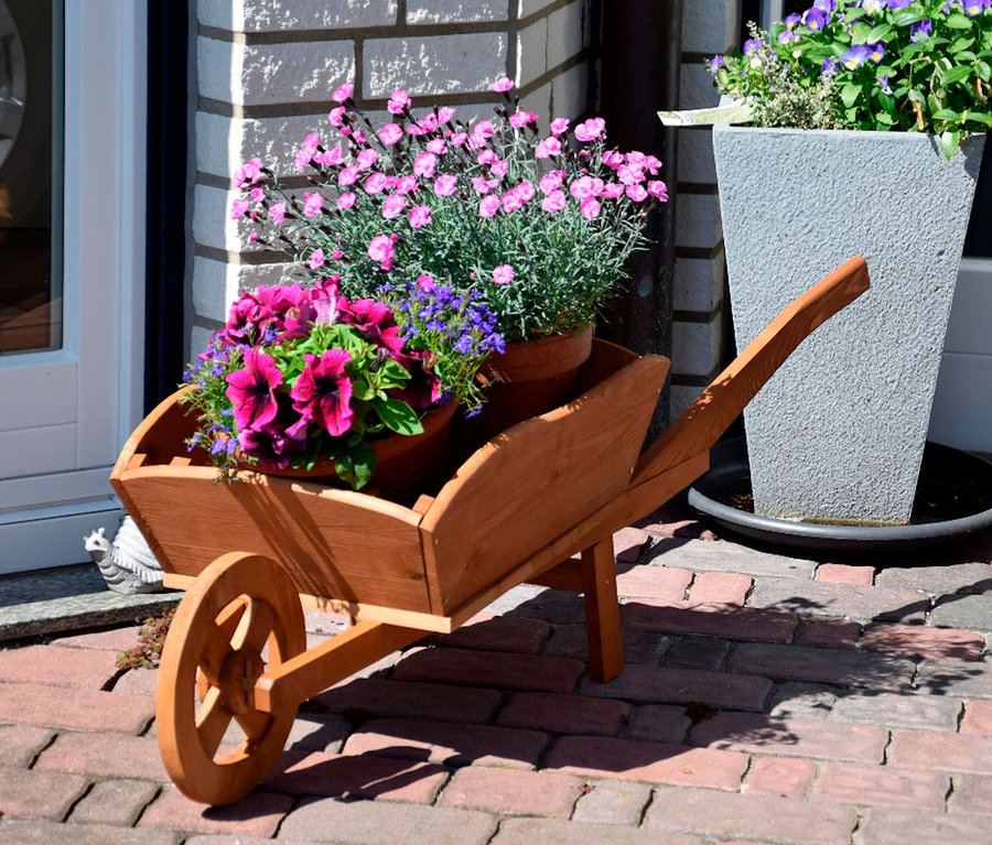 Eine Pflanzschubkarre XXL aus Holz mit Blumen steht auf einer Ziegelterrasse.