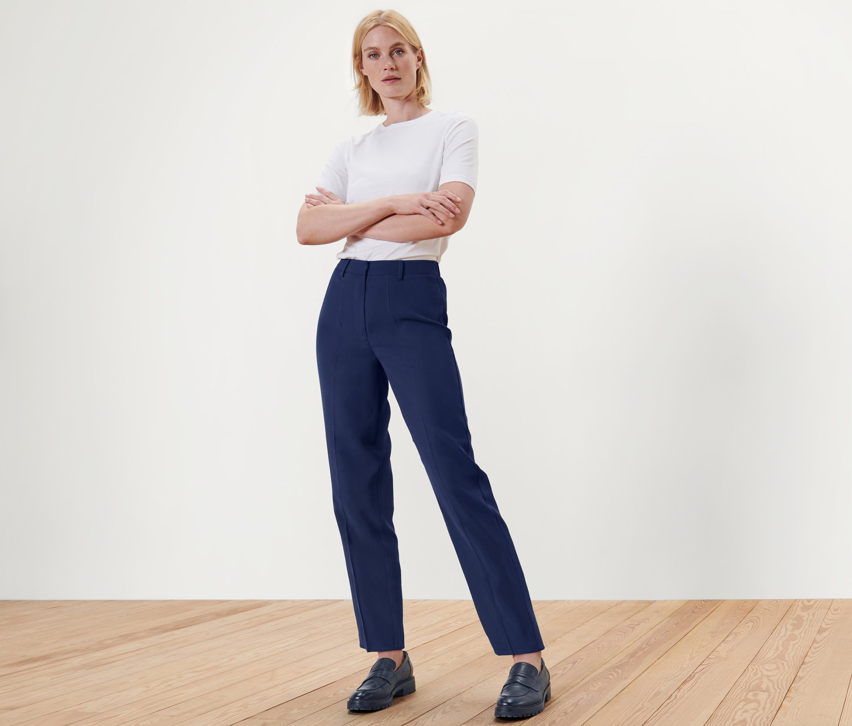 Blonde Frau steht mit verschränkten Armen in blauen Webhosen und Schuhen auf Holzboden.