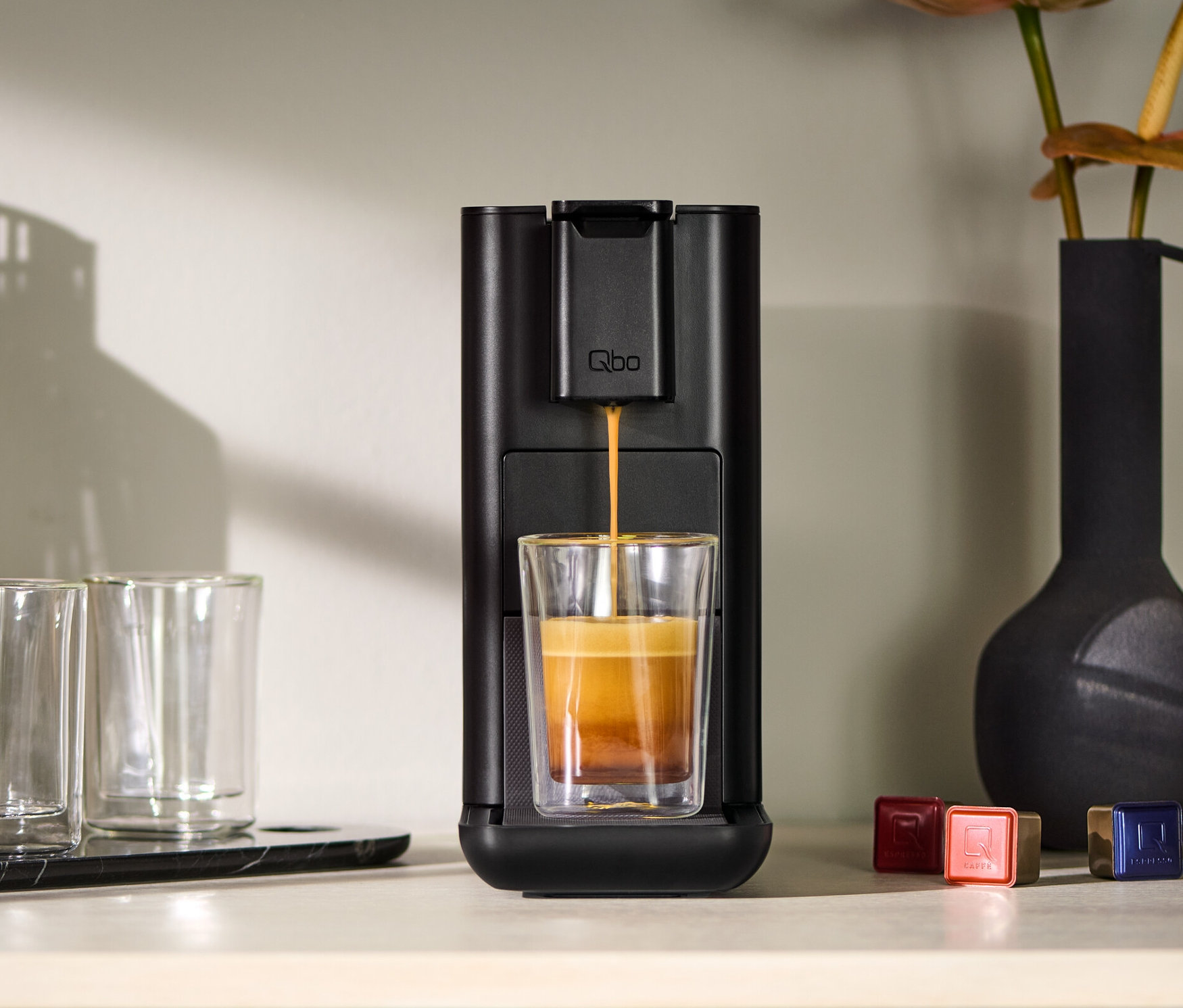 Qbo ICONIC Dark Chrome Kaffeemaschine füllt ein Glas mit Kaffee. Daneben stehen zwei leere Gläser und eine Vase.