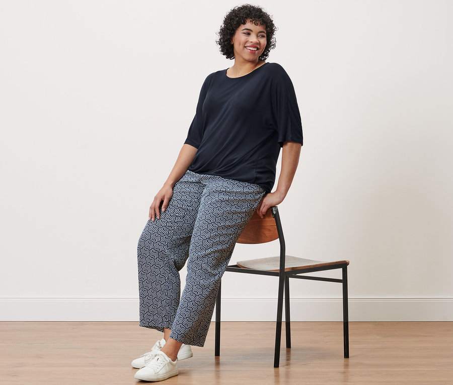 Frau sitzt auf einem Stuhl und trägt ein dunkelblaues Blusenshirt und eine Webhose mit Alloverprint.
