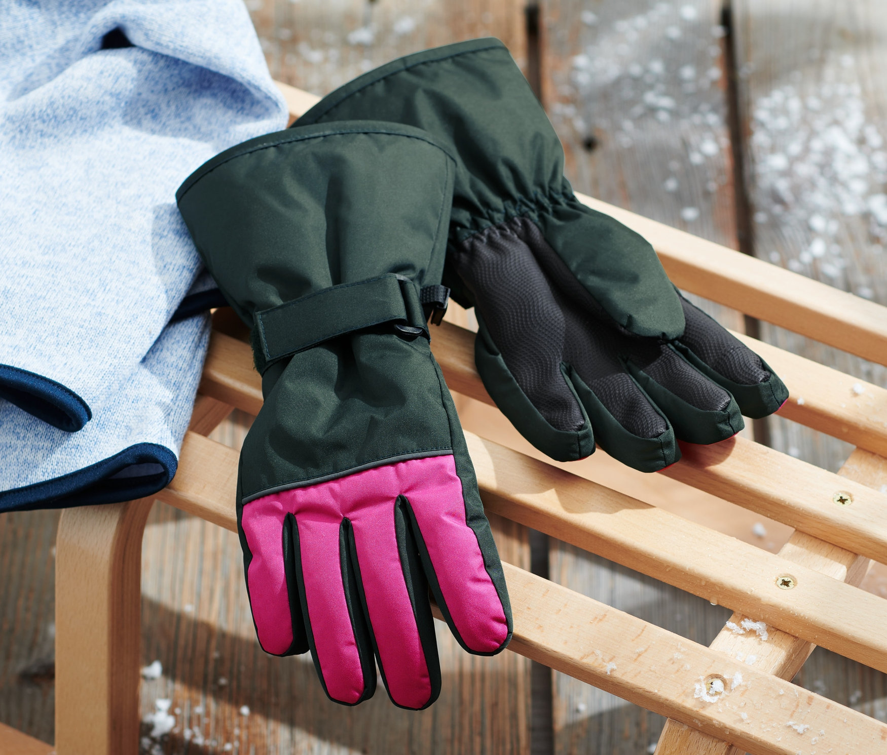 Pinke Kinder-Ski- und Winterhandschuhe liegen neben einem Pullover auf einem Holzschlitten.