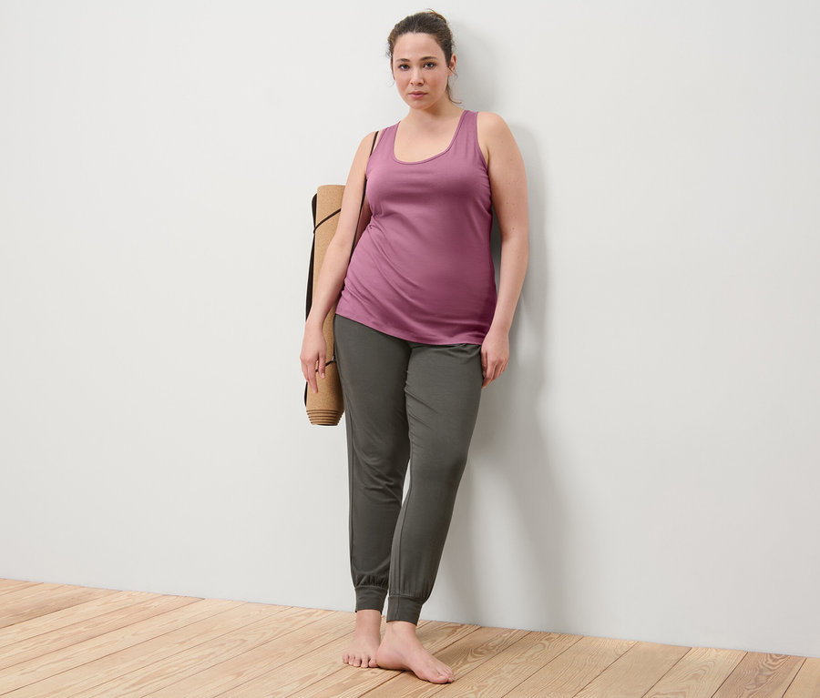 Frau lehnt an Wand, trägt 2 Tops in Mauve und Yogahose, eine Yogamatte über der Schulter.