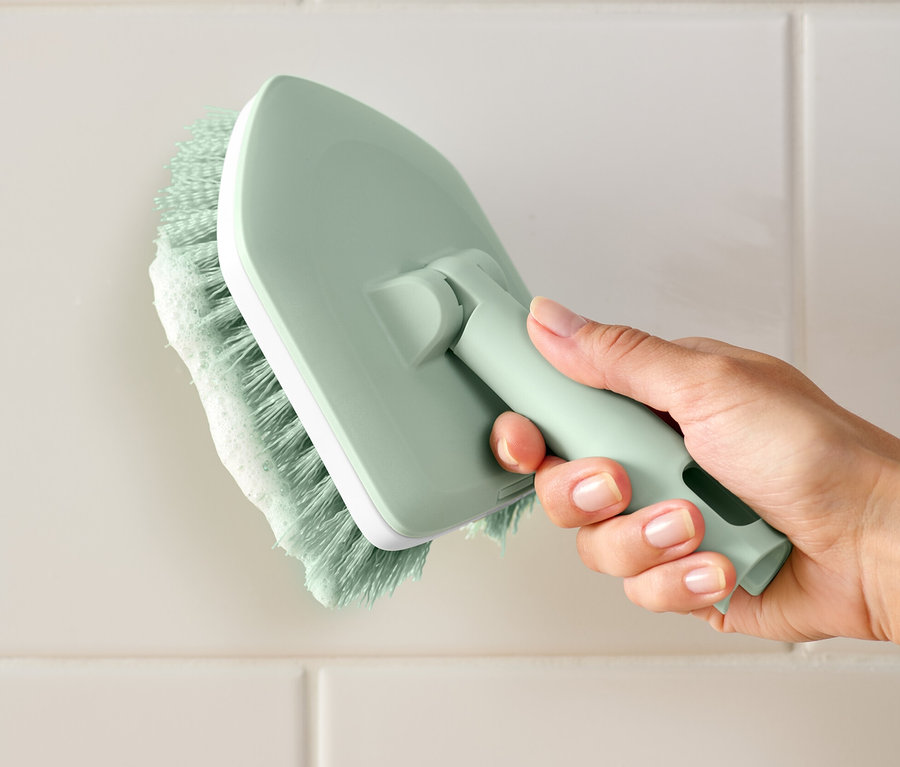 Eine Hand hält einen Fliesen-und-Badschrubber und reinigt weiße Fliesen.