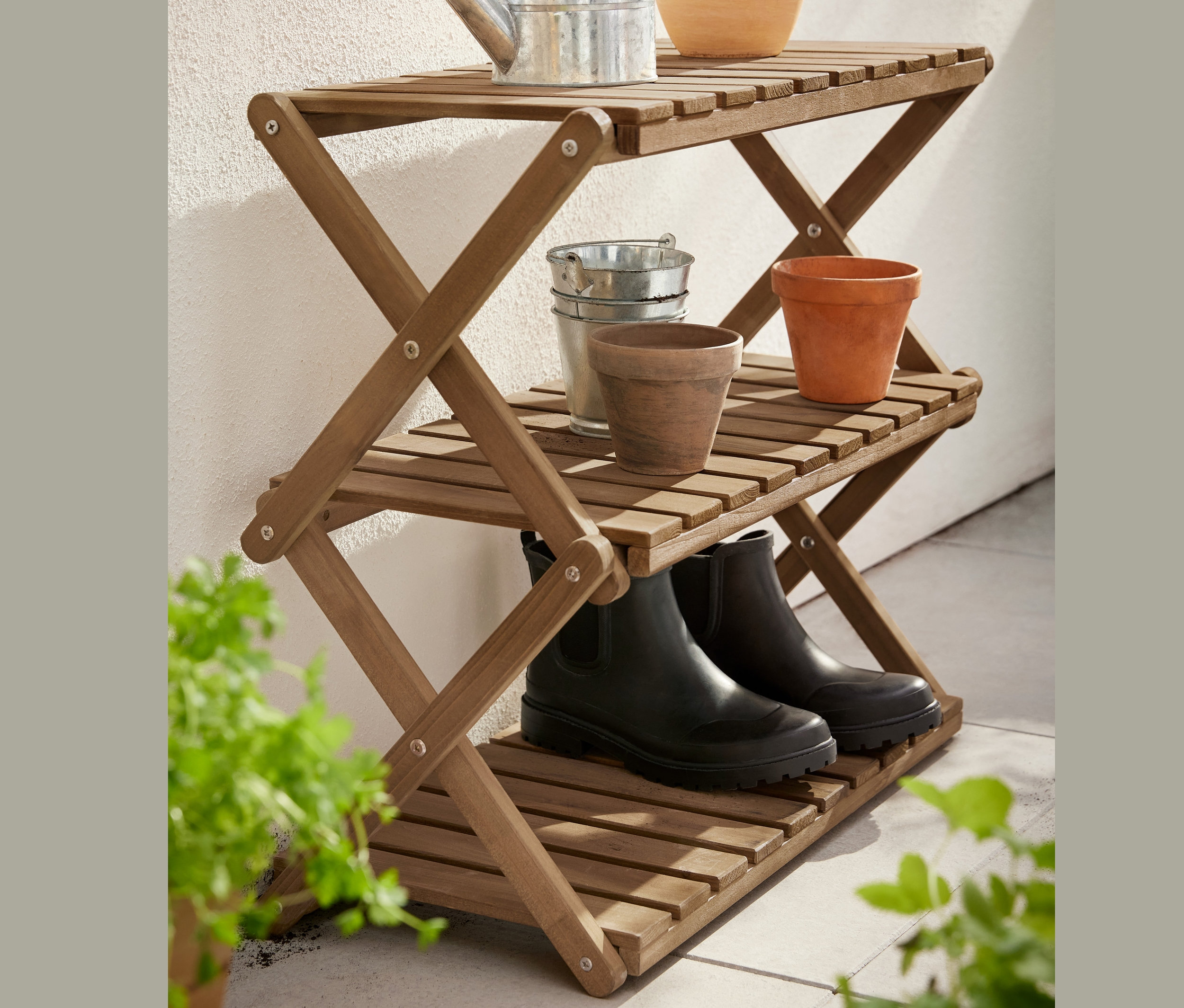 Auf den drei Ablagen des zusammenklappbaren Regals »Tinus« befinden sich Blumentöpfe, eine Gießkanne und Gummistiefel.