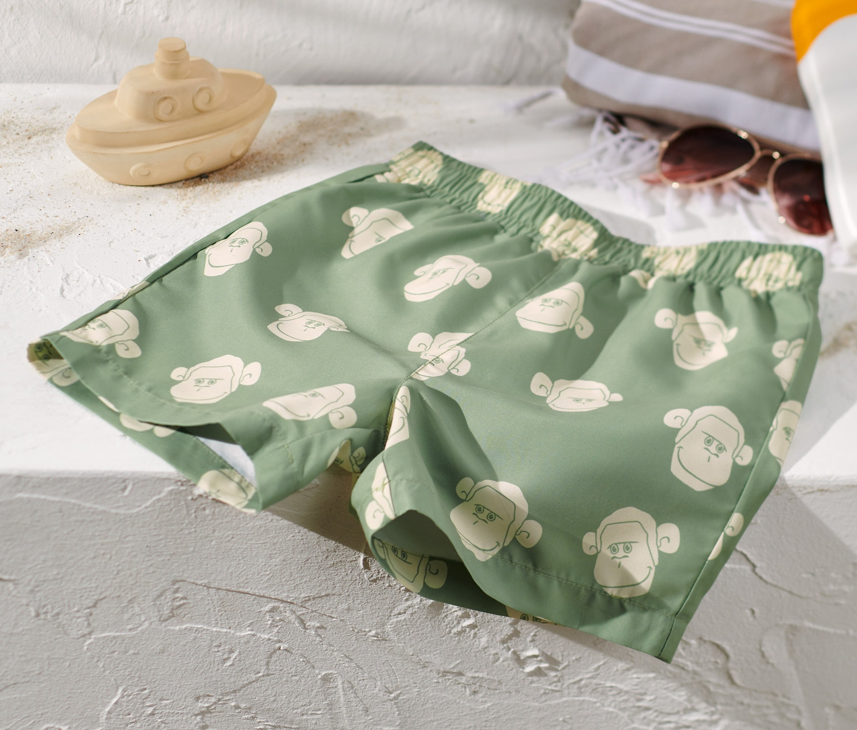 Grüne Kinder-Boardshorts mit Affenmuster liegen auf einer weißen Oberfläche neben einem Spielzeugboot, einem Handtuch, einer Sonnenbrille und Sonnencreme.