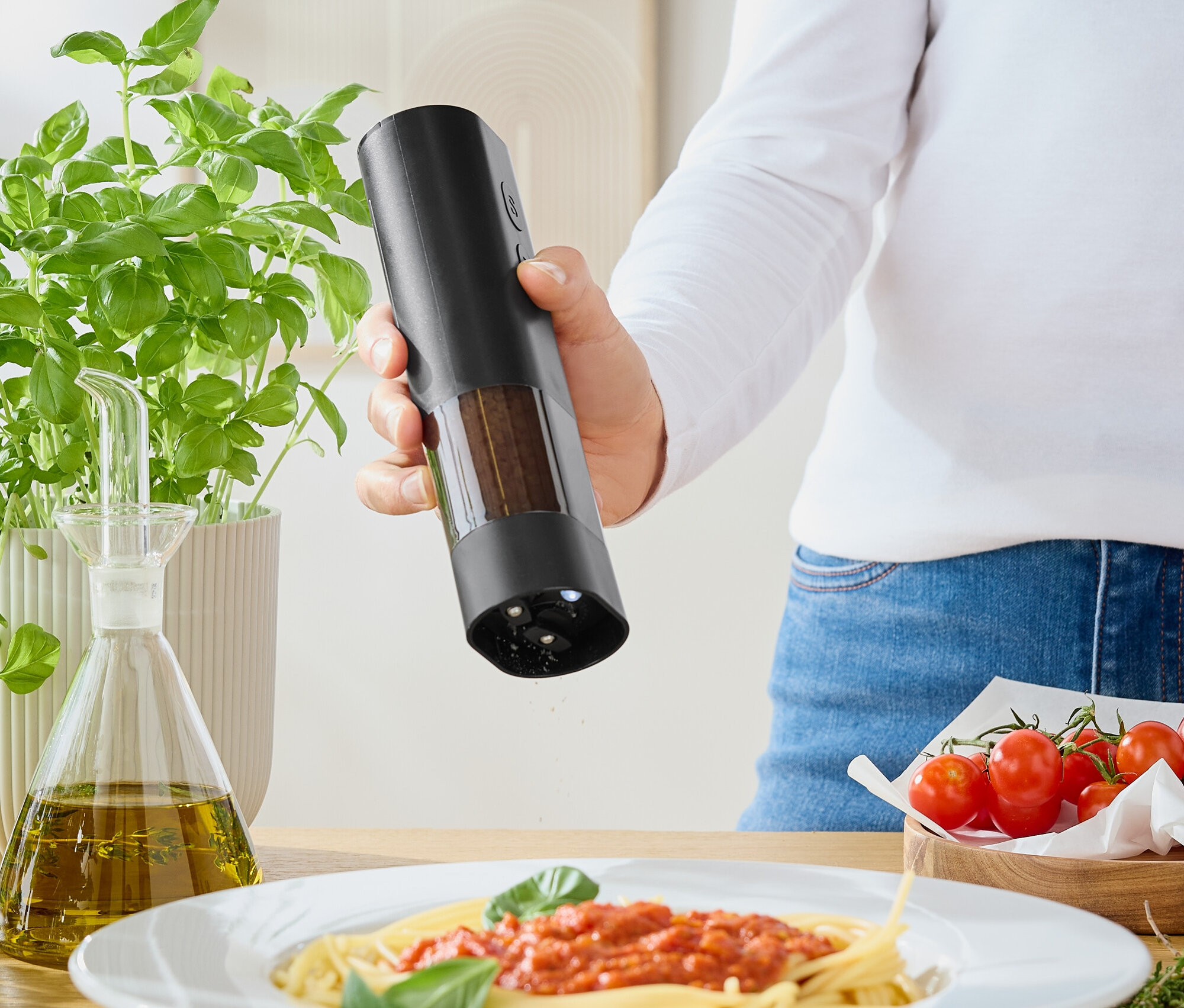 Frau würzt Spaghetti mit Tomatensoße mit einer Mühle. Auf dem Tisch stehen Basilikum, Olivenöl und Tomaten.