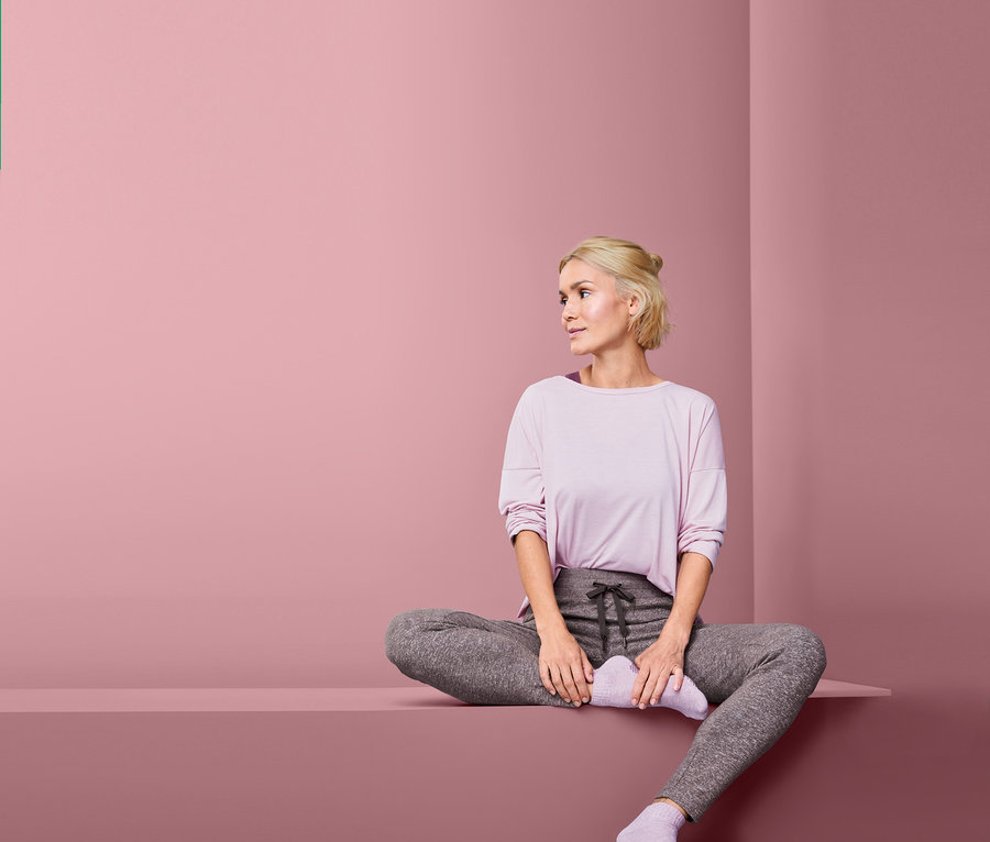 Blonde Frau sitzt mit Schneidersitz auf einem Podest und trägt eine Relaxhose.