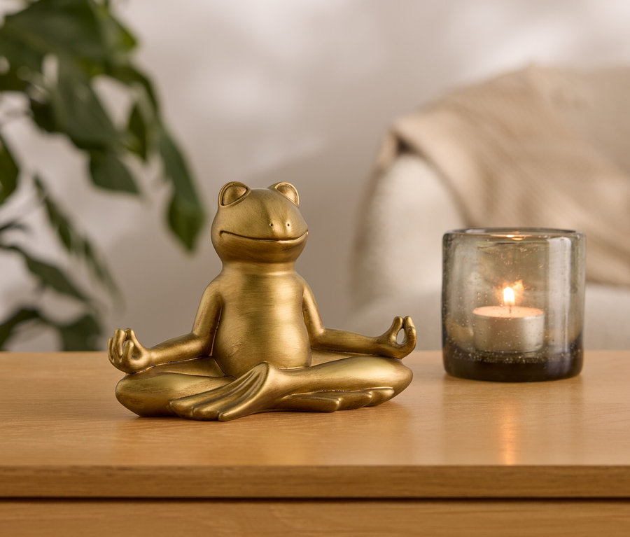 Eine goldene Froschfigur sitzt in einer Meditationshaltung neben einem Glasgefäß mit einer brennenden Kerze.