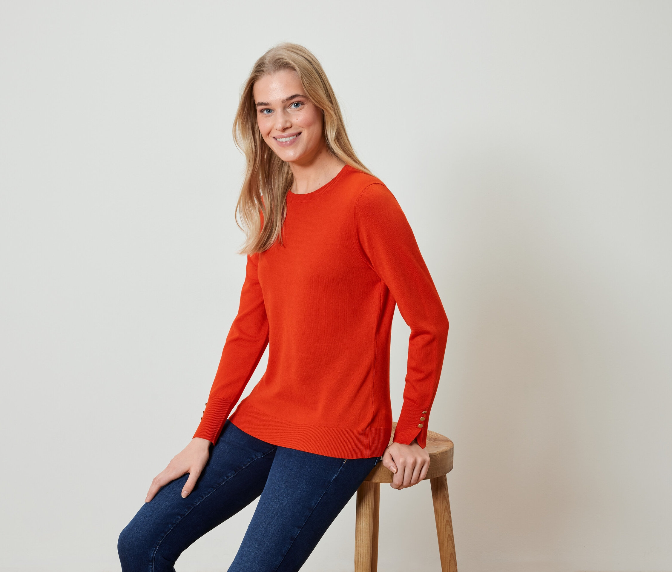 Blonde Frau sitzt auf einem Hocker und trägt einen orangefarbenen Pullover und blaue Jeans.