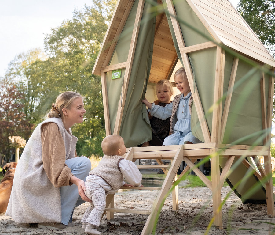 Eine Frau hält ein Kind, das auf eine Leiter zum EXIT Spielhaus »Yuki 300« mit Rutsche klettert, im Inneren sind zwei Mädchen.