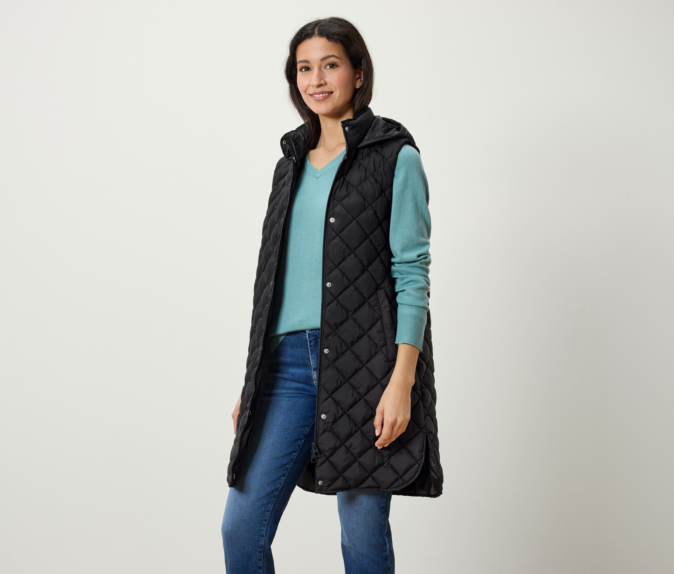 Eine Frau trägt eine lange, gesteppte, schwarze Weste mit Kapuze, einen blauen Pullover und Jeans.