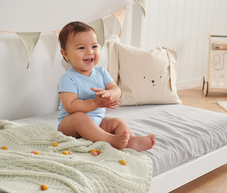 Lächelndes Baby sitzt in einem blauen 3er-Pack Bodys aus Bio-Baumwolle auf einem Bett.