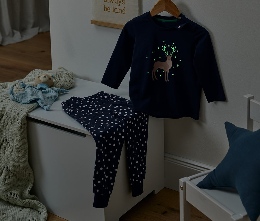 Dunkelblauer Glow-in-the-dark-Pyjama mit Bio-Baumwolle und Hirschmotiv auf einem Bügel, Hose mit weißen Punkten liegt auf einer Truhe.