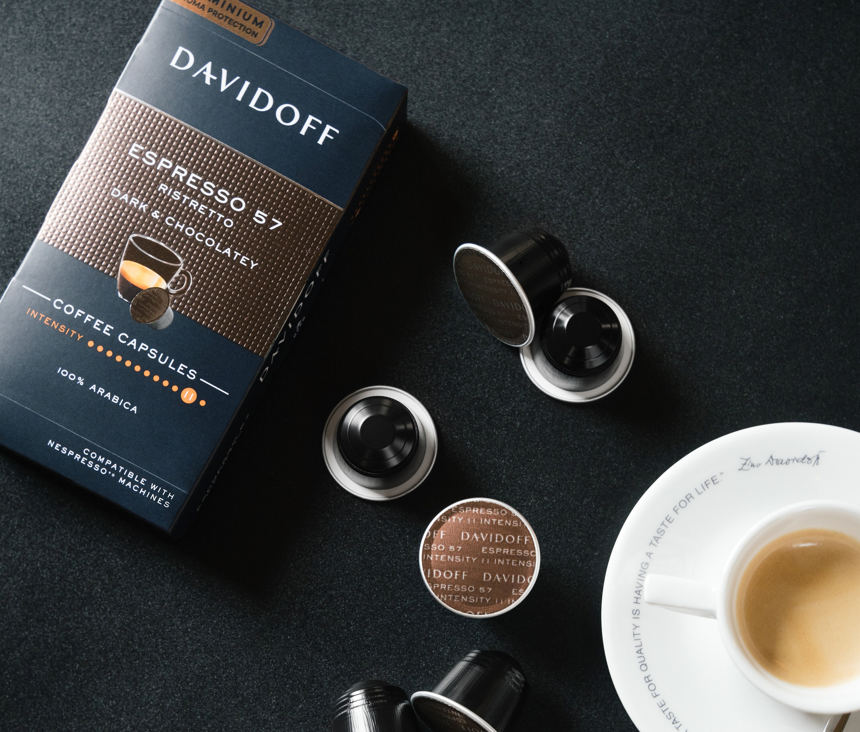 Eine Packung und mehrere Kapseln DAVIDOFF CAFÉ Espresso 57 Ristretto neben einer Tasse Espresso auf schwarzem Hintergrund.