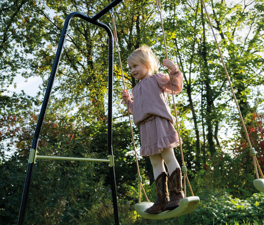 Mädchen in Kleid und Cowboystiefeln steht auf einer EXIT Doppelschaukel »Ivy«.