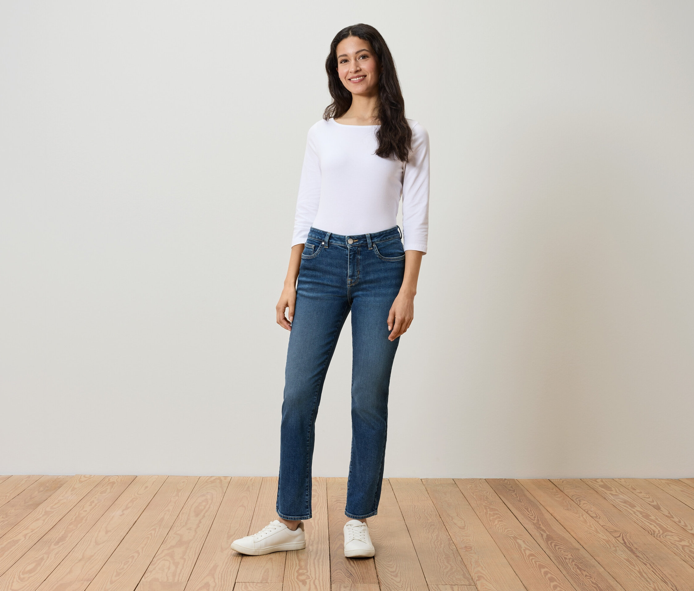 Eine Frau steht auf einem Holzboden und trägt eine weiße Dreiviertelarmbluse, blaue Jeans und weiße Turnschuhe.