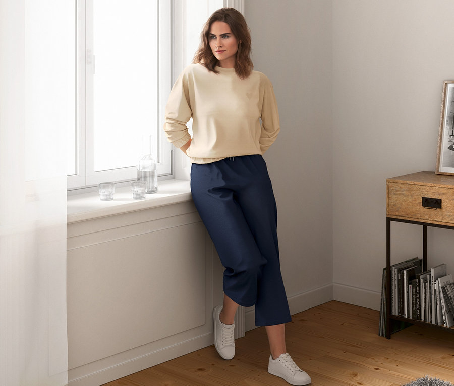 Frau lehnt am Fensterbrett, trägt beiges Sweatshirt und blaue Culottes.