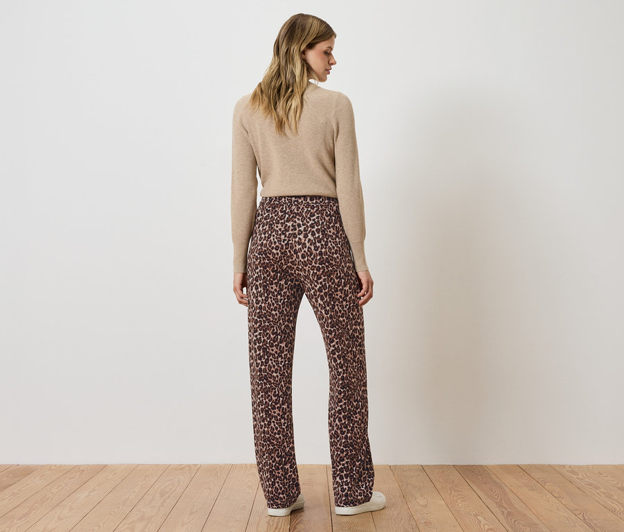 Frau steht mit dem Rücken zur Kamera und trägt einen beigen Pullover und eine braune Hose mit Tiermuster.