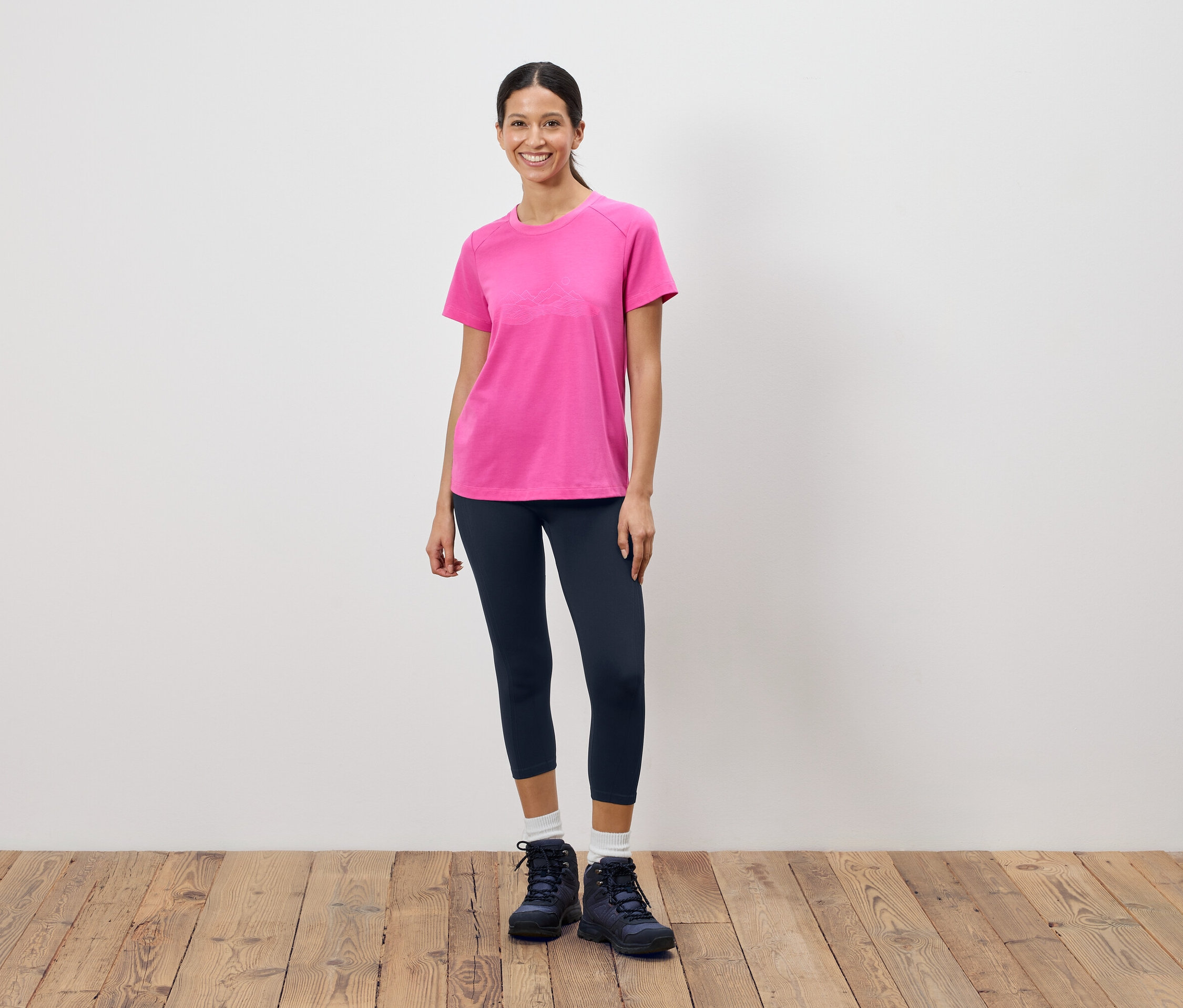 Frau steht vor einer weißen Wand und trägt ein pinkes T-Shirt, dunkle Leggings und Wanderschuhe.