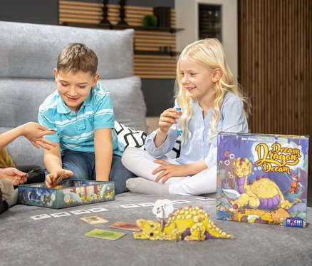 Drei Kinder sitzen auf einem grauen Sofa und spielen das Spiel „Dream Dragon Dream“.