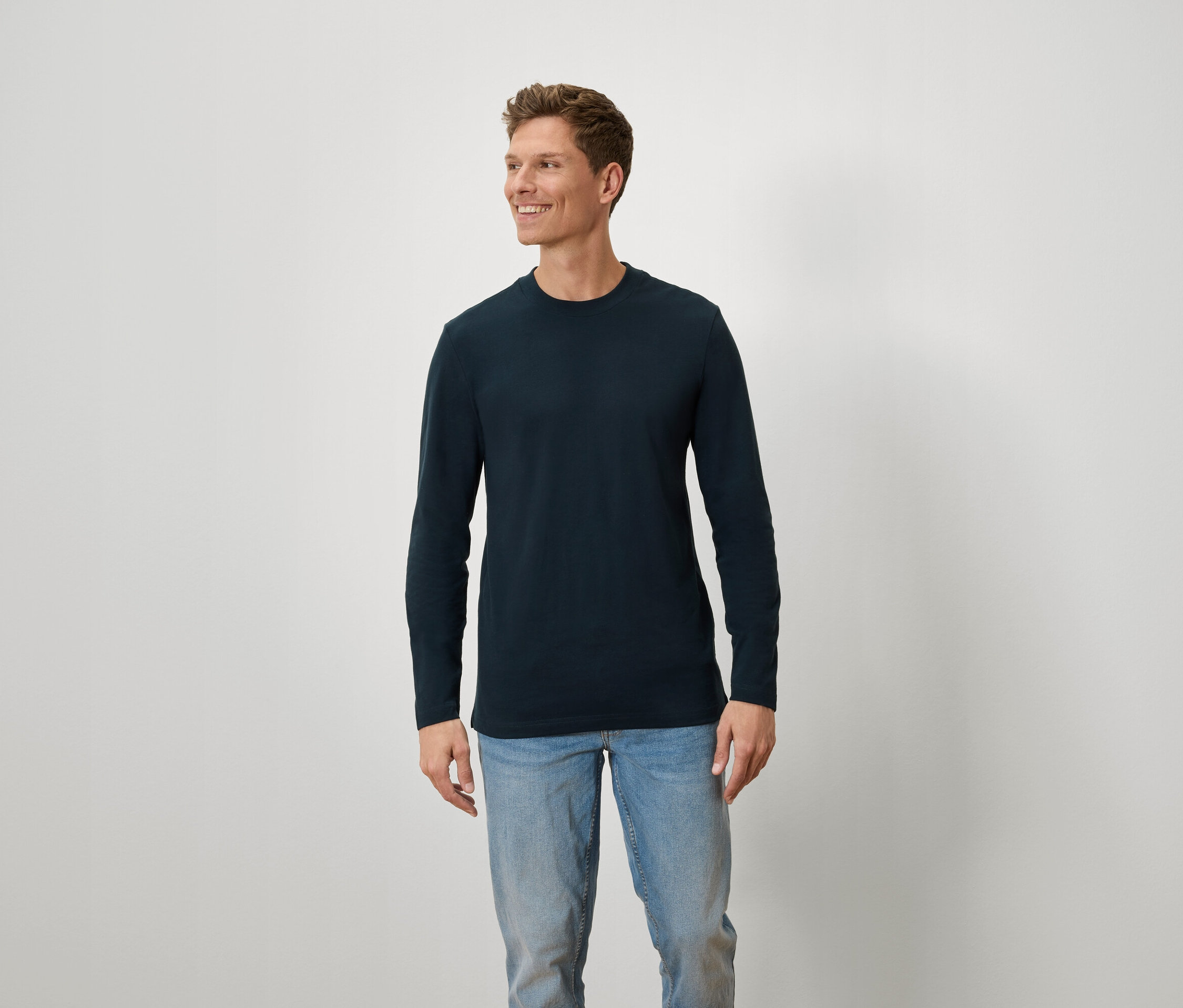 Mann mit braunen Haaren und blauen Augen trägt ein blaues Langarmshirt und Jeans.